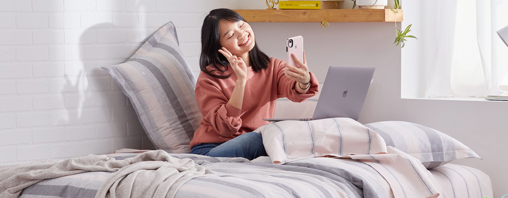 College student sitting on bed with phone and laptop.