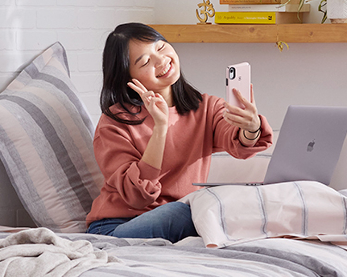 College student sitting on bed with phone and laptop.